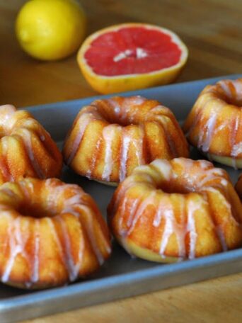 Six mini bundt cakes with white glaze sit on a metal tray. In the background, there are whole lemons, an orange, and a halved pink grapefruit on a wooden surface.