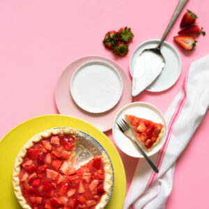 Strawberry pie on yellow cake stand with plates and pie servers on pink surface