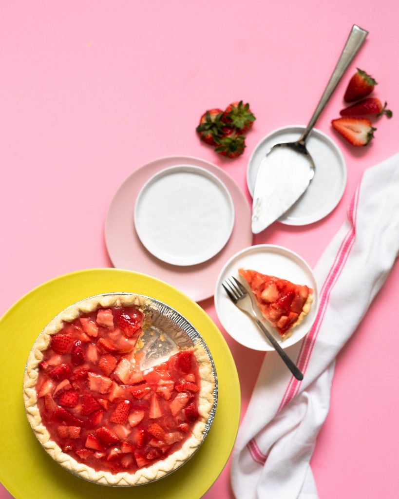 Strawberry pie on yellow cake stand with plates and pie servers on pink surface