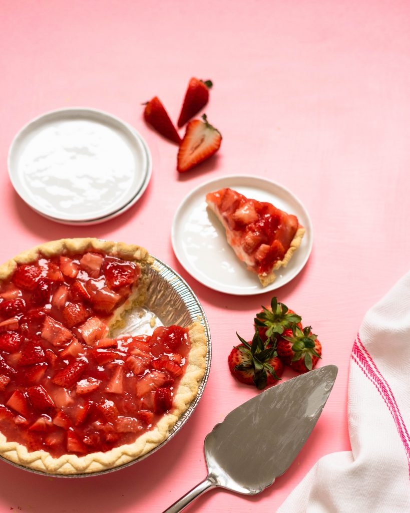 Strawberry pie, slice of strawberry pie on a plate, and stack of plates on pink surface