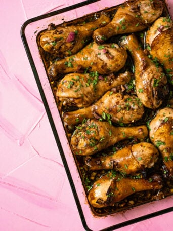 A baking tray filled with glazed balsamic chicken legs, garnished with chopped fresh herbs, set on a textured pink surface.
