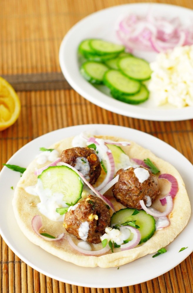 White plate with pita topped with Greek meatballs, tzatziki, red onion, and cucumber on bamboo placemat.