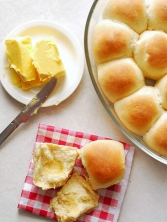 A dish of golden brown dinner rolls sits beside a plate with sliced butter and a butter knife. Two dinner rolls, one split open with butter, rest on a red and white checkered napkin.