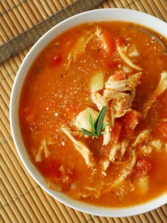 A bowl of tomato-based chicken soup with shredded chicken, carrots, tomatoes, potatoes, and a sprig of rosemary sits on a bamboo mat—perfect for anyone who enjoys hearty fare like turkey and lentil soup.