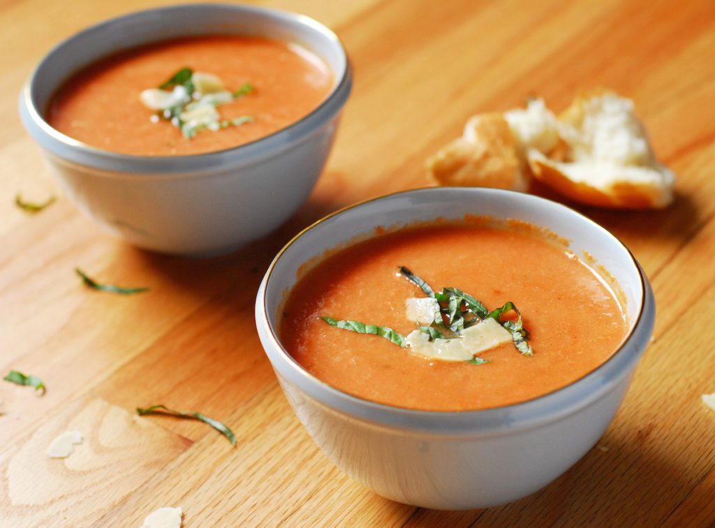 Bowls of tomato soup on wooden surface with hunks of bread and spoons