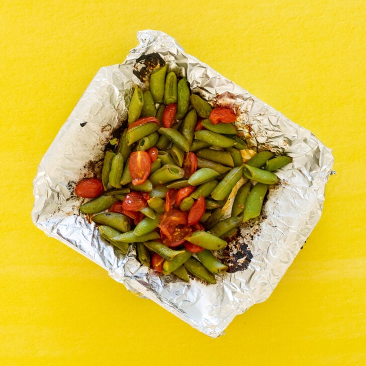 A foil packet filled with roasted green beans and cherry tomatoes sits on a bright yellow surface, perfect alongside a fresh grilled corn salad.