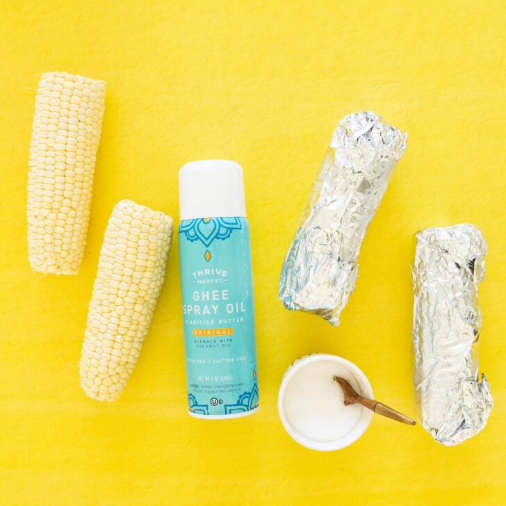 Three ears of raw corn, two wrapped in foil, a can of ghee spray oil, and a small bowl of salt with a wooden spoon are arranged on a bright yellow background—perfect ingredients for a fresh grilled corn salad.