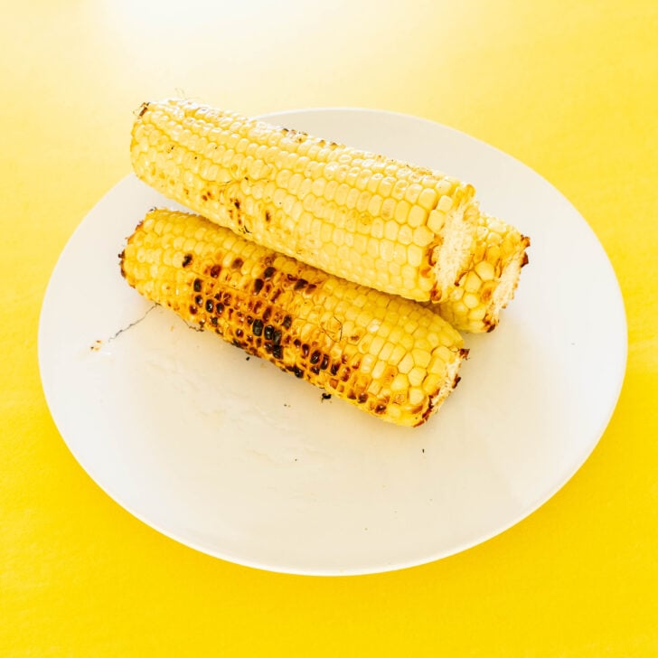 Two grilled corn on the cob pieces are stacked on a white plate, set against a bright yellow background—perfect for inspiring your next grilled corn salad.