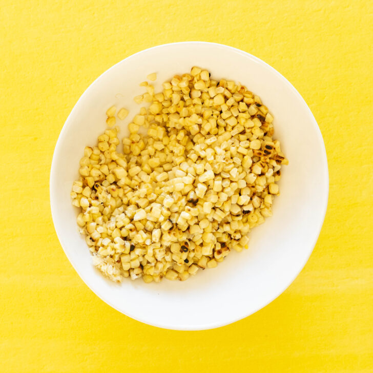 A white bowl filled with roasted corn kernels sits on a bright yellow surface, ready to be tossed into a flavorful grilled corn salad.