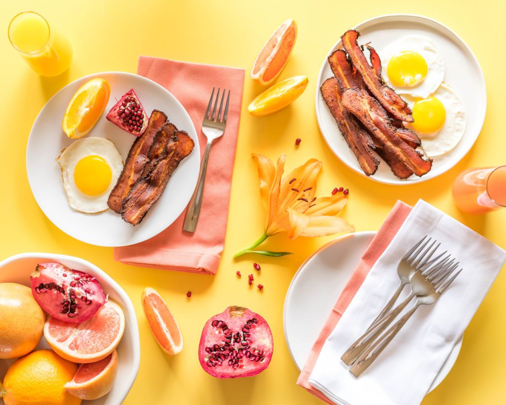 Sous Vide Bacon, eggs, and fruit arranged on white plates and a yellow background