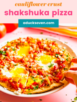 Cauliflower crust shakshuka pizza on a white rounded plate with a hand reaching for a slice.