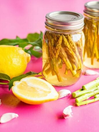 Two jars of sous vide pickled asparagus sit on a pink surface next to a whole lemon, a halved lemon, fresh asparagus spears, green leaves, and garlic cloves.