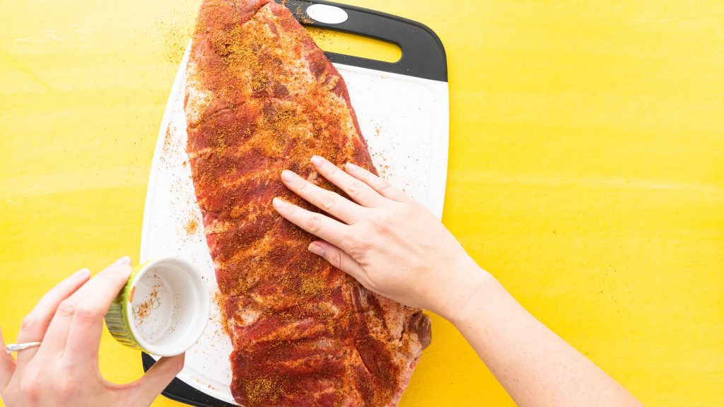 Rack of ribs on white cutting board on yellow surface being rubbed with seasoning