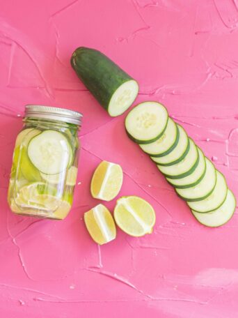 A mason jar of sous vide infused tequila with cucumber and lemon slices sits on a pink textured surface. Next to it are a halved cucumber, cucumber slices, and four lemon wedges arranged neatly.