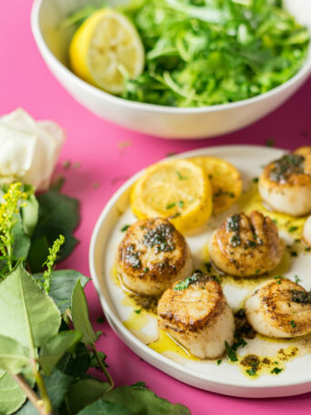 A plate of sous vide scallops garnished with herbs and lemon slices, with a bowl of fresh green salad and a halved lemon in the background, on a pink surface adorned with white flowers and greenery.