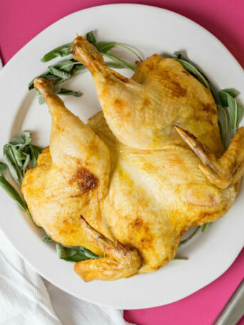 A beautifully roasted spatchcock chicken sits on a white plate with green herbs, set on a pink surface beside a white napkin, three whole lemons, a fork, and a knife.