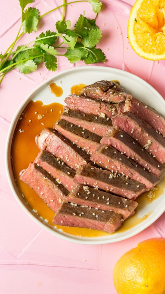 Orange Marinated Sous Vide Strip Steak on a white plate, arranged on a pink background
