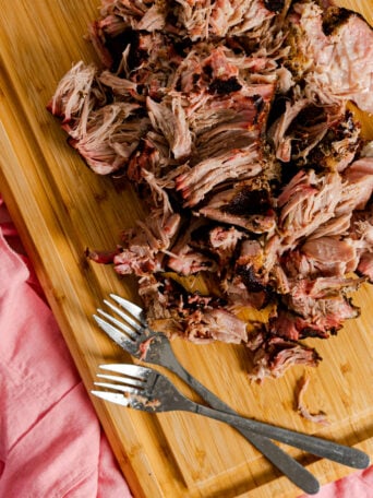 Sous vide smoked pork shoulder is shredded and piled on a wooden cutting board next to two metal forks, with a pink cloth partially visible to the side.