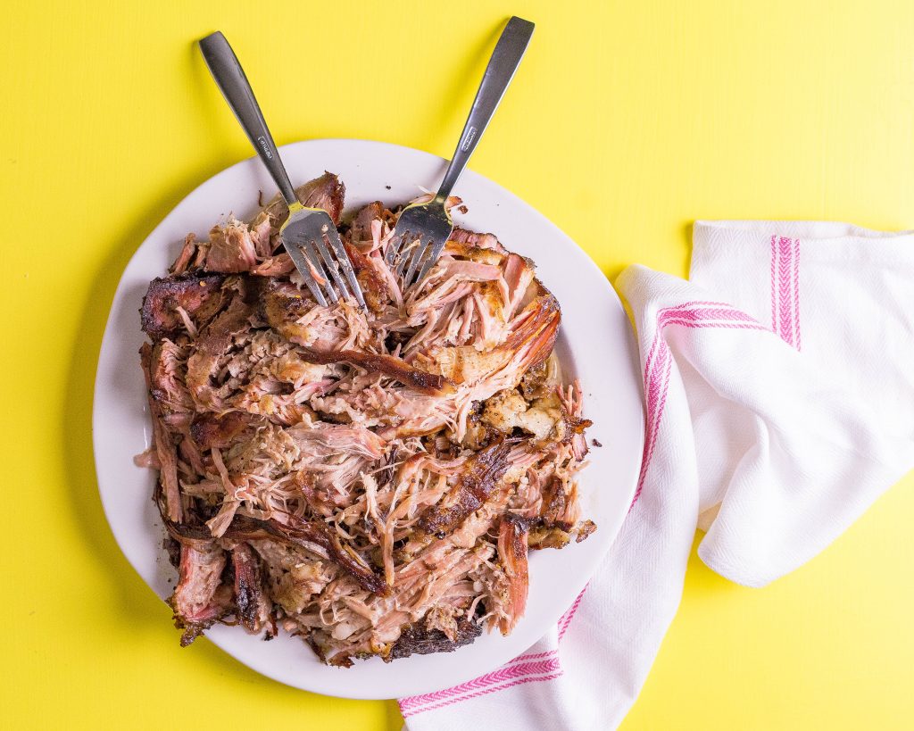 Shredded sous vide pork shoulder on a white plate with two forks, on a yellow background with a white cloth.