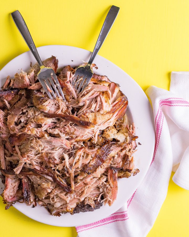 Shredded pork shoulder on a white plate with two forks, on a yellow background with a white cloth.