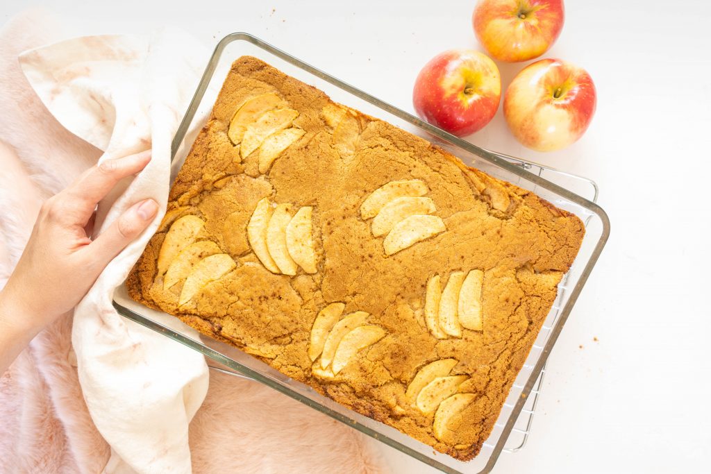 Brown butter apple blondies in glass baking dish being set down on white surface with apples