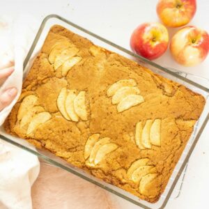 Brown butter apple blondies in glass baking dish being set down on white surface with apples