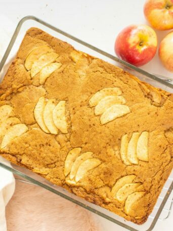 Brown butter apple blondies in glass baking dish being set down on white surface with apples