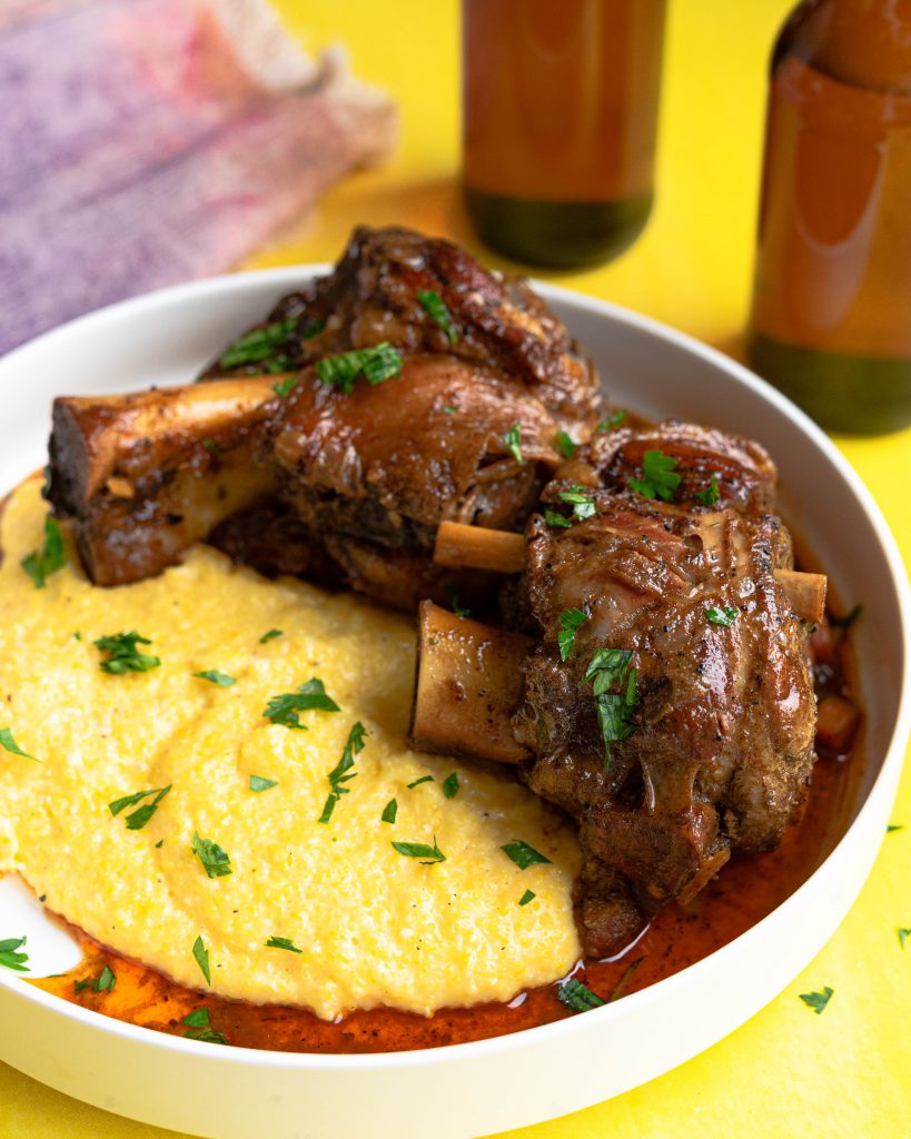 Braised pork shanks over grits on a white platter on a yellow background