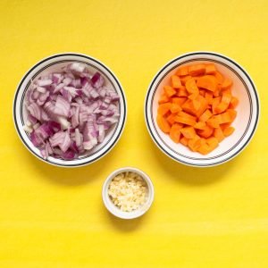 Red onions, carrots, and minced garlic in bowls on yellow surface