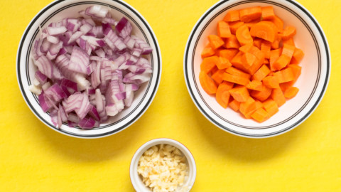 Three bowls on a yellow surface: one with chopped red onions, one with chopped carrots, and a small bowl in the center with minced garlic—perfect aromatics for preparing flavorful Braised Pork Shanks.