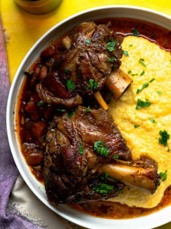 A bowl with two braised pork shanks in sauce served alongside creamy polenta, garnished with chopped herbs. The bowl sits on a bright yellow surface with a purple napkin nearby.
