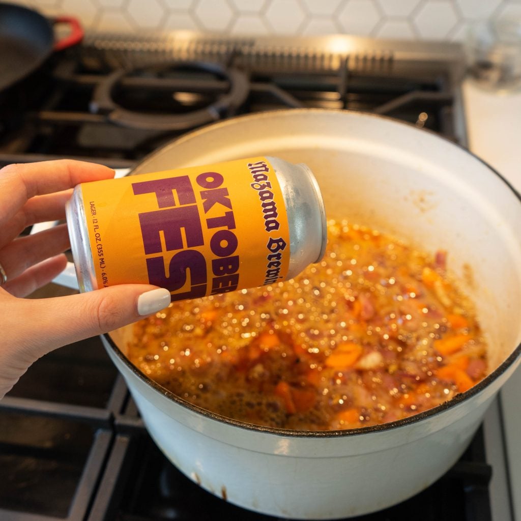 A hand holds a can of Oktoberfest beer above a white pot on a stove, pouring it into a bubbling mixture of chopped vegetables and liquid—perfect for preparing Braised Pork Shanks.