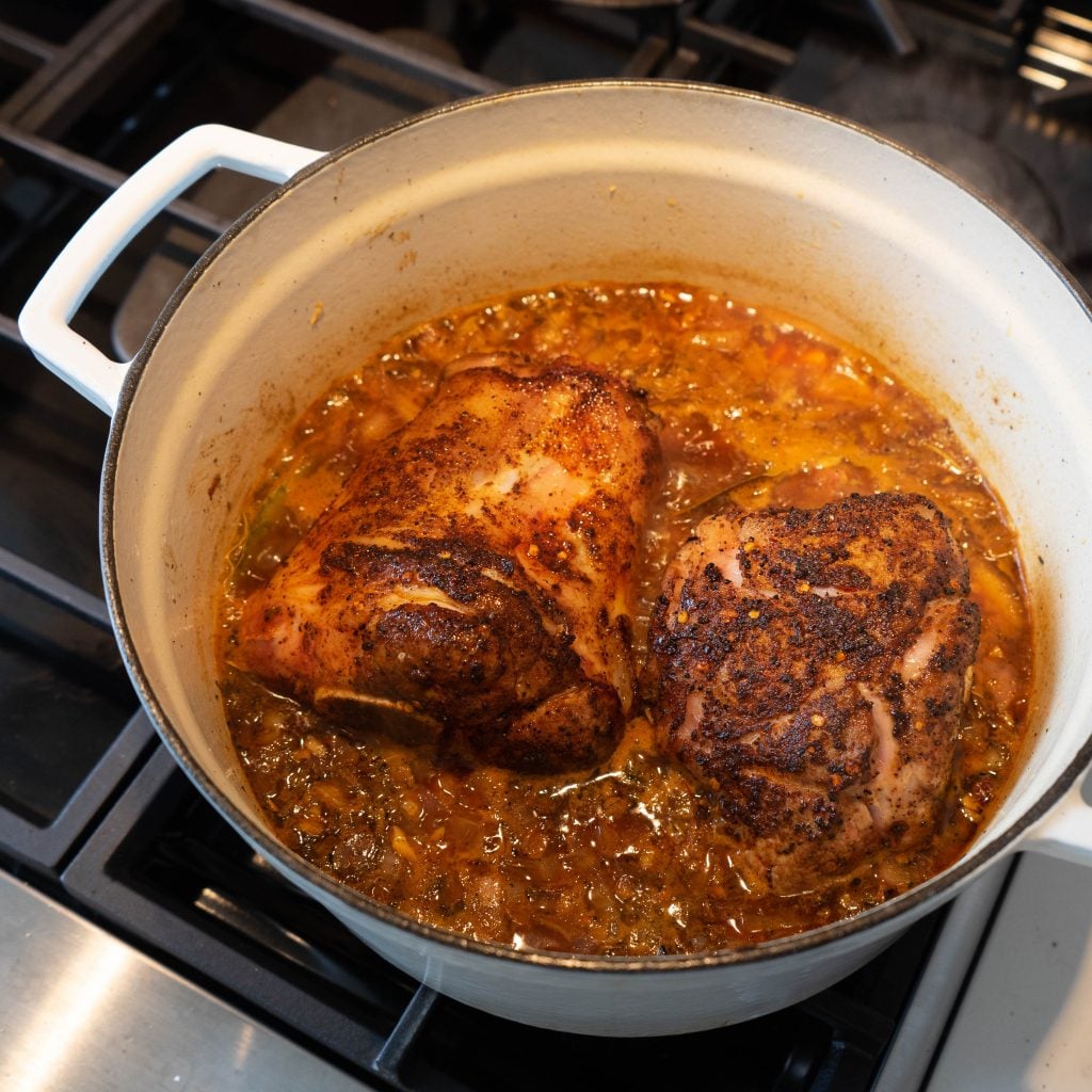 Two seasoned Braised Pork Shanks are simmering in a Dutch oven filled with a rich, chunky tomato and herb sauce. The white pot with handles sits on the stovetop, capturing the essence of slow-cooked comfort food.