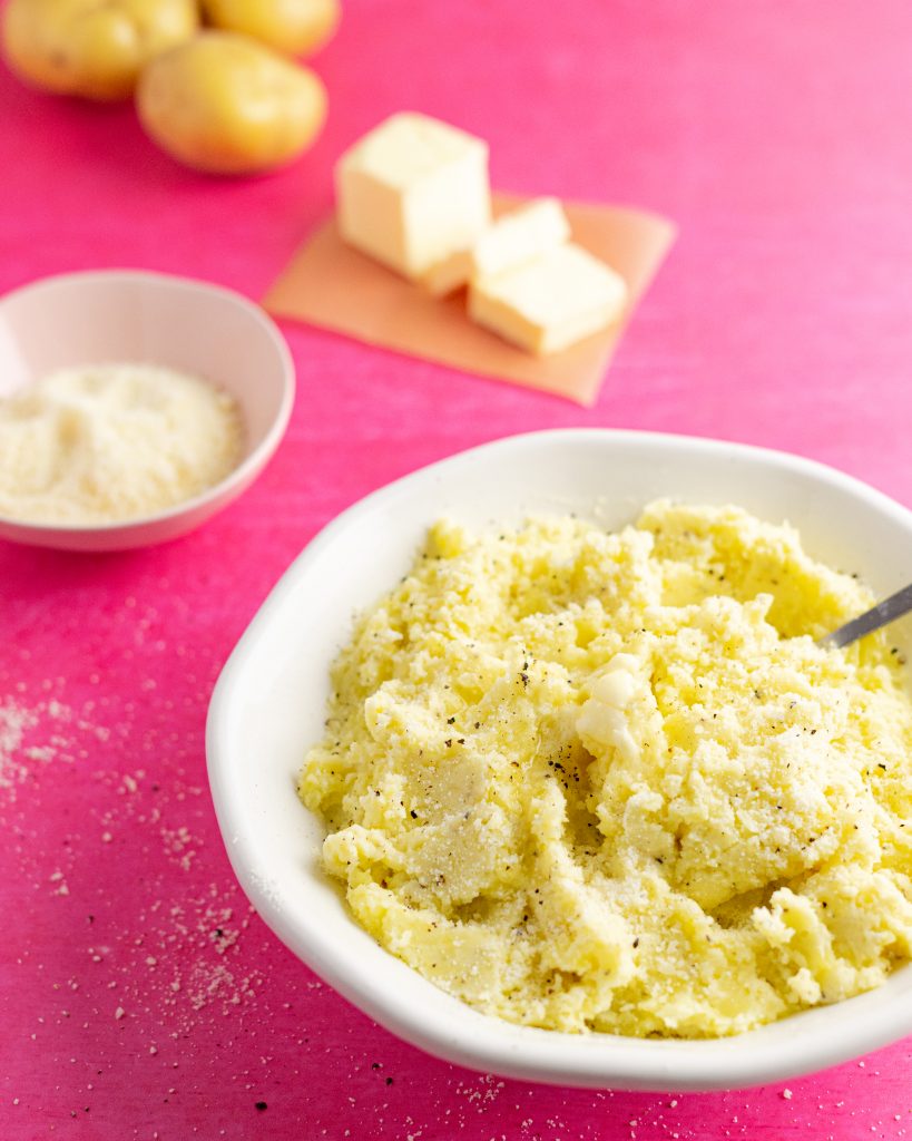 Bowl of mashed potatoes on hot pink background with bowl of parmesan, butter, and whole potatoes