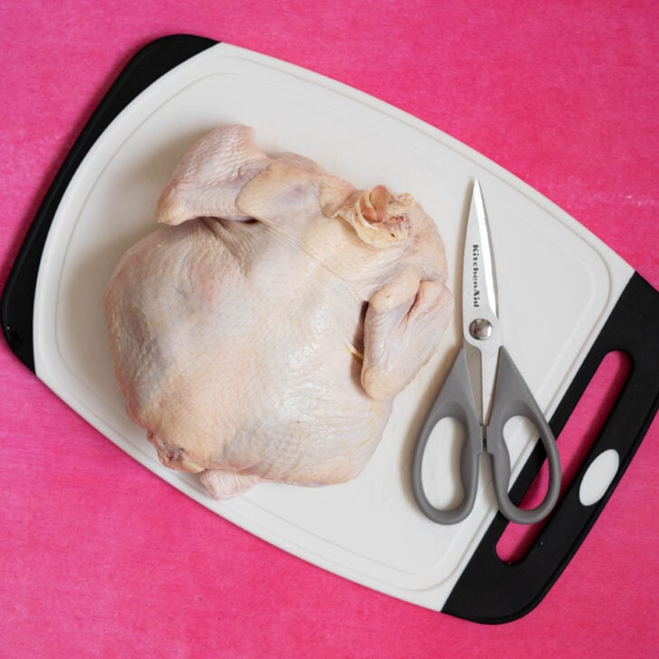 A sous vide whole chicken and a pair of kitchen scissors are placed on a white cutting board with a black edge, set against a bright pink background.