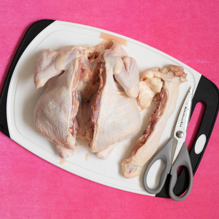 A raw sous vide whole chicken, partially cut open with poultry shears, sits on a white cutting board with a black handle, all placed on a bright pink surface.
