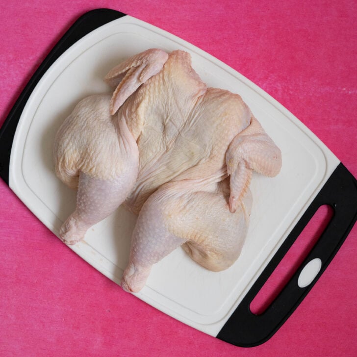 A raw, spatchcocked chicken, ready for a sous vide whole chicken recipe, rests on a white cutting board with black edges against a bright pink background.