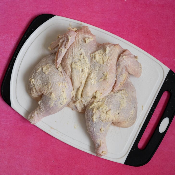 Raw, butterflied sous vide whole chicken coated with a butter and herb mixture sits on a white cutting board with a black handle, placed on a bright pink surface.