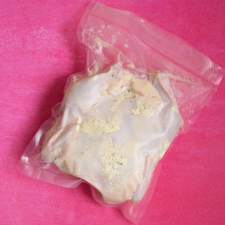 A sous vide whole chicken, sealed in a vacuum plastic bag and coated with seasoning, rests on a bright pink background.