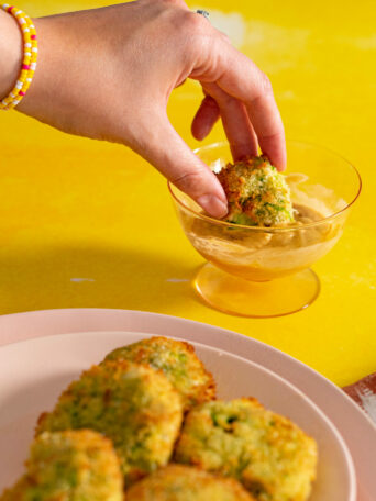 Hand dipping air fried zucchini fritter into dipping sauce with yellow background