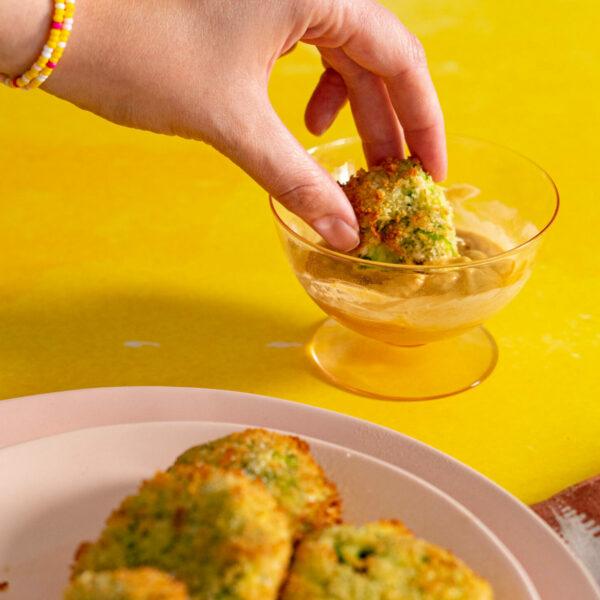 Hand dipping air fried zucchini fritter into dipping sauce with yellow background