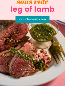 Sliced Sous Vide boneless leg of lamb with a drizzle of salsa verde on top on a white plate with a fork on the side and a bowl of salsa verde.