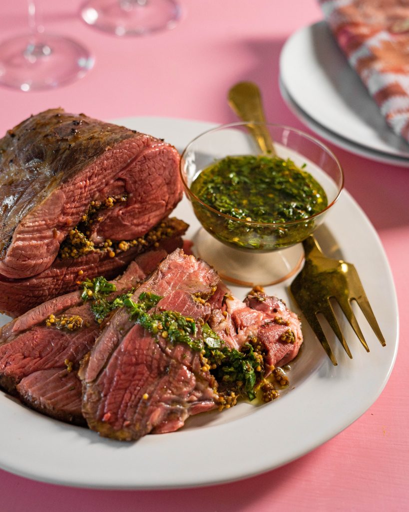 Leg of lamb on serving platter and sliced with salsa verde on pink background.