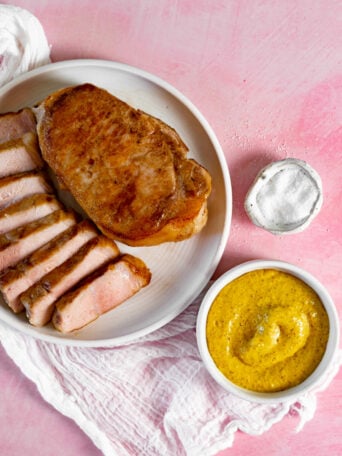 A plate with a cooked sous vide pork chop, half sliced, sits on a pink surface next to a small bowl of yellow mustard sauce and a small dish of salt, with a white cloth underneath the plate.