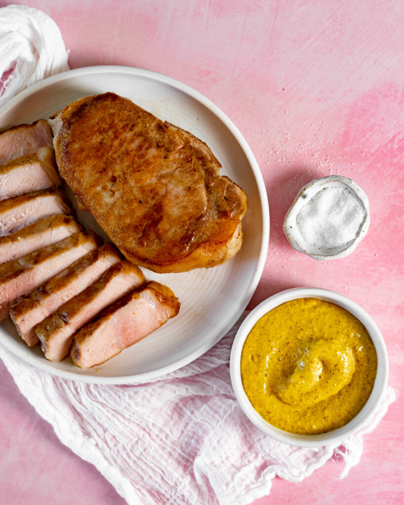 Sliced pork chop and whole pork chop on white plate with a bowl of mustard and bowl of salt on pink background