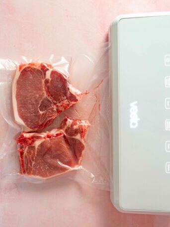 Two raw sous vide pork chops are vacuum-sealed in a plastic bag next to a white vacuum sealer machine on a light pink surface.