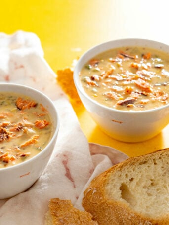Two white bowls filled with creamy salmon chowder topped with shredded vegetables sit on a light cloth, next to slices of crusty bread, all on a bright yellow surface.