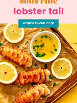 Sous Vide Lobster Tail garnished with parsley on parchment paper on top of a cookie sheet. Lemon wedges are by the lobster along with garlic butter sauce in a white bowl.