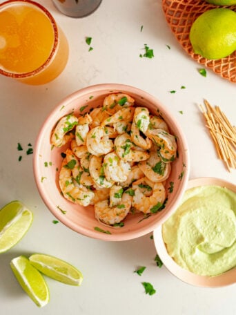 A bowl of tender sous vide shrimp garnished with chopped herbs sits beside lime wedges, a creamy green dip, wooden skewers, a lime, and a glass of orange beverage on a white surface.