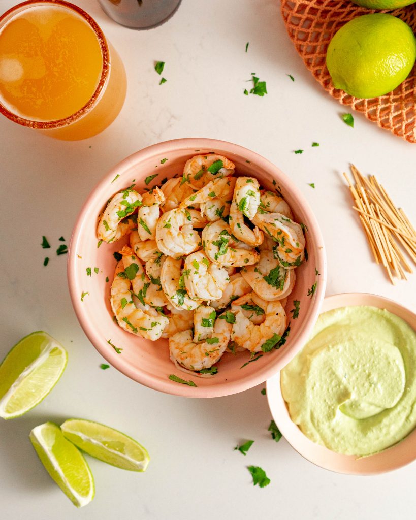 Shrimp in pink bowl with bowl of avocado lime cream sauce next to it on white surface.